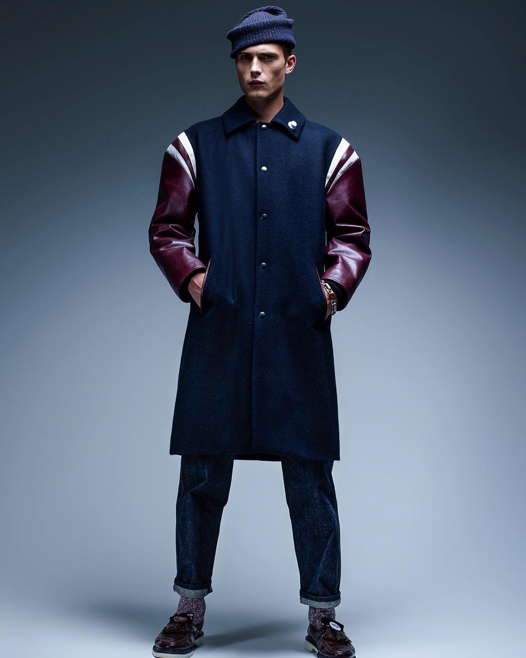 Full-length shot of a male model in a navy coat with leather sleeves, dark jeans, and a knit hat, posing against a grey background.