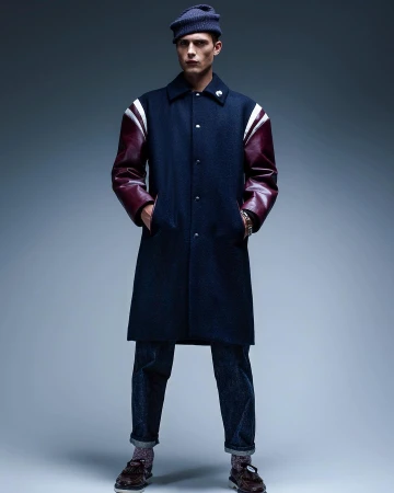 Full-length shot of a male model in a navy coat with leather sleeves, dark jeans, and a knit hat, posing against a grey background.