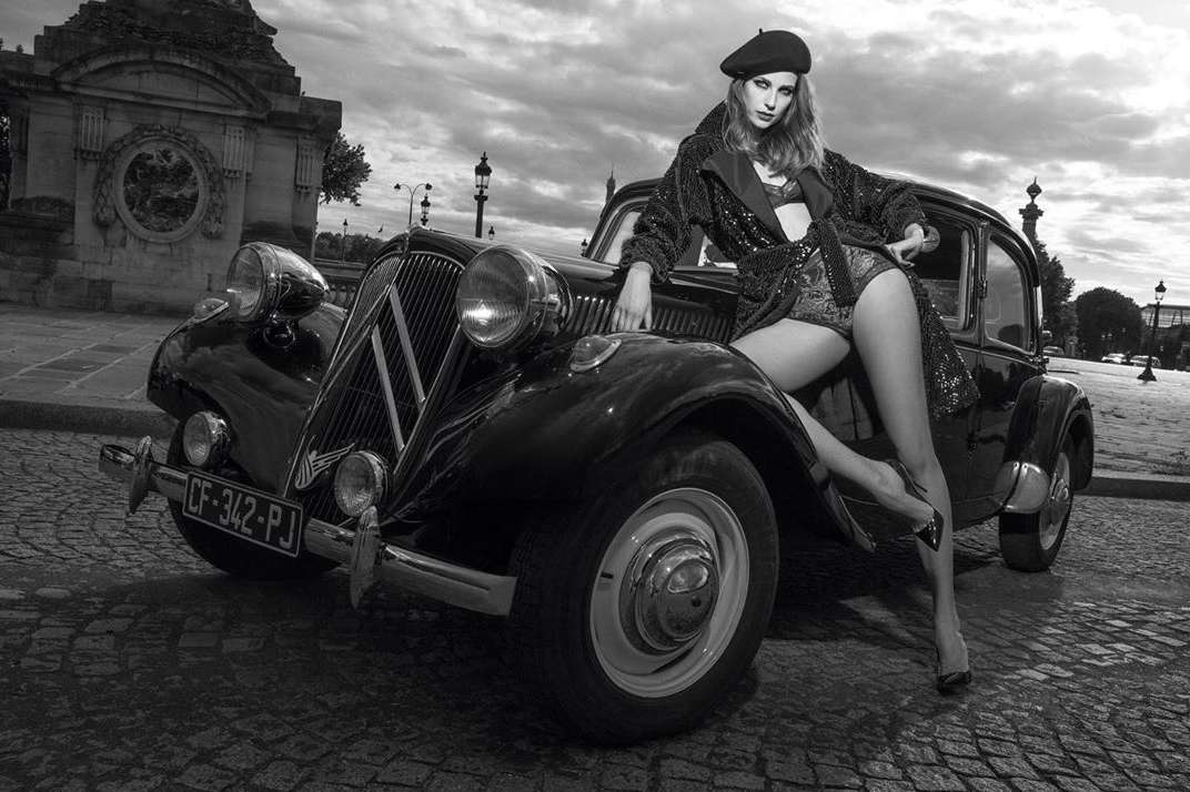 Monochrome fashion shot of a woman in a beret and sequined outfit, posing on a vintage car in front of an ornate stone structure.