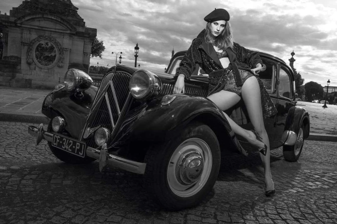 Monochrome fashion shot of a woman in a beret and sequined outfit, posing on a vintage car in front of an ornate stone structure.