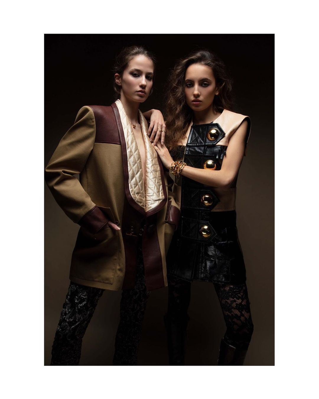 Two young women in fashionable outfits pose indoors in a portrait-style image. One wears a beige jacket and the other a black and beige vest.