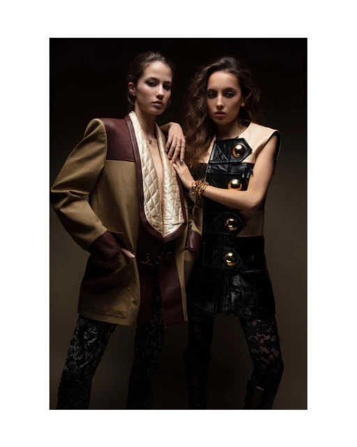 Two young women in fashionable outfits pose indoors in a portrait-style image. One wears a beige jacket and the other a black and beige vest.