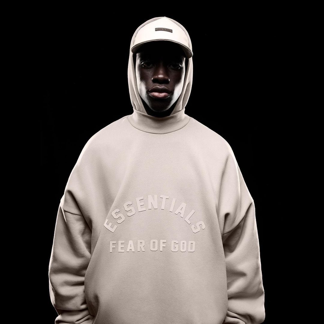 A dark-skinned model poses in a photo studio wearing a beige Essentials by Fear of God sweatshirt and baseball cap against a black background.