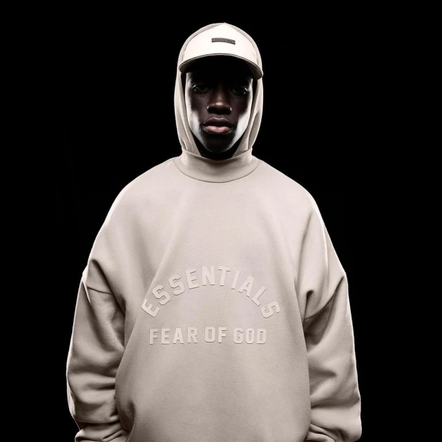 A dark-skinned model poses in a photo studio wearing a beige Essentials by Fear of God sweatshirt and baseball cap against a black background.