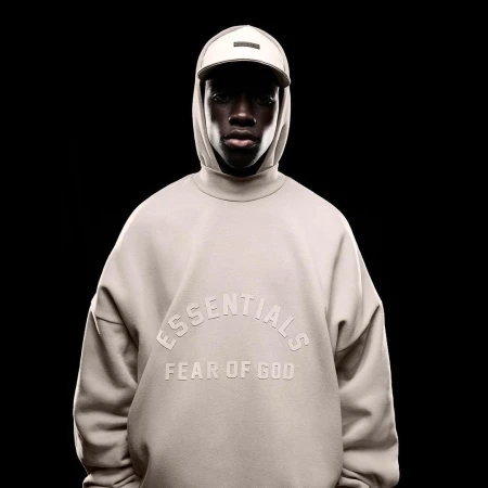 A dark-skinned model poses in a photo studio wearing a beige Essentials by Fear of God sweatshirt and baseball cap against a black background.