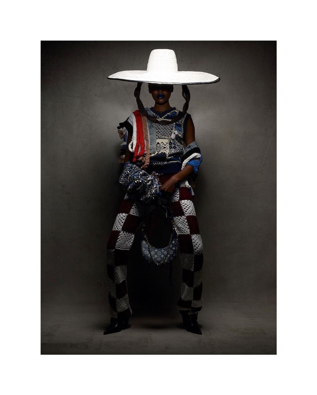 Fashionable person in checkered pants, knit top, and large hat in a studio shot, showcasing unique textures and design elements.