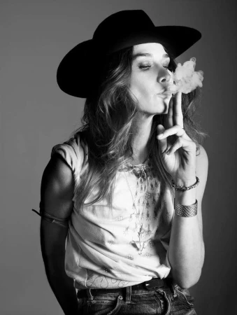 Monochrome portrait of a woman wearing a wide-brimmed black hat and light-colored top, blowing smoke from her fingers.