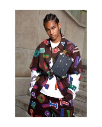 Young man wearing a brown printed shirt and shorts with a black crossbody bag standing outdoors against stone steps.