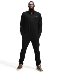 A man wearing a black utility jumpsuit and brown sneakers posing against a solid white background.