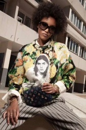 A Black woman with voluminous hair wearing large sunglasses, a patterned sweater, and holding a small dark handbag outdoors.