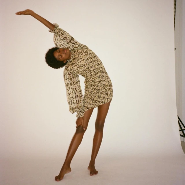 An adult Black female model in a printed mini dress poses in a studio with her arm raised against a white backdrop, showcasing fashion and style.