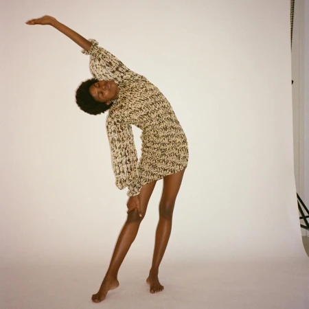 An adult Black female model in a printed mini dress poses in a studio with her arm raised against a white backdrop, showcasing fashion and style.