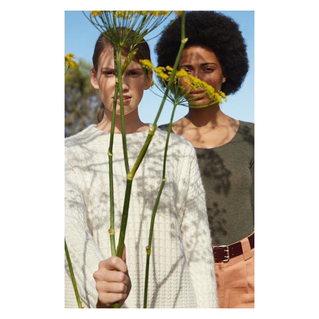 Two women pose in an outdoor setting, partially obscured by stems and yellow flowers. One wears a textured sweater, the other an olive shirt.