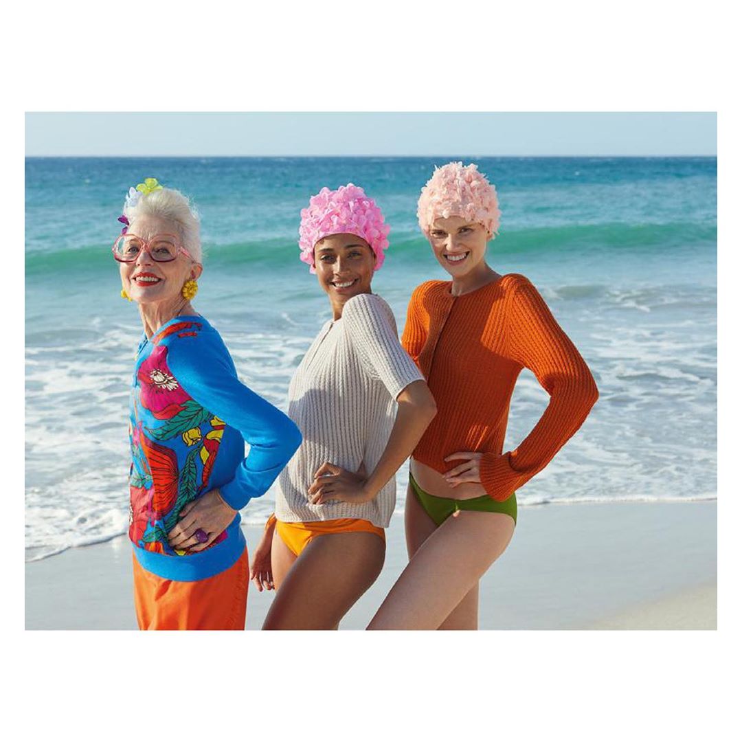 Three women in swim caps pose on a sandy beach in bright sweaters and swimwear, smiling against an ocean backdrop.