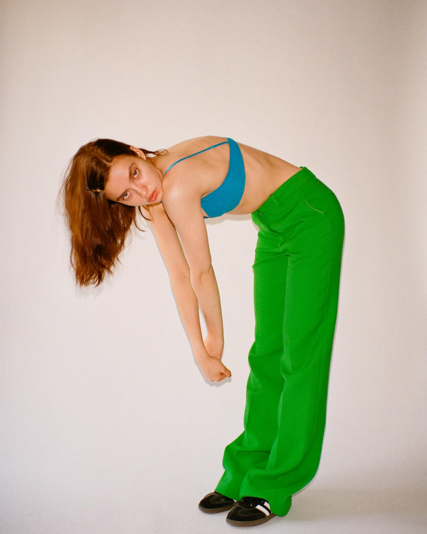 An adult woman with long brown hair in green pants and a blue top poses with a forward bend against a plain white background.