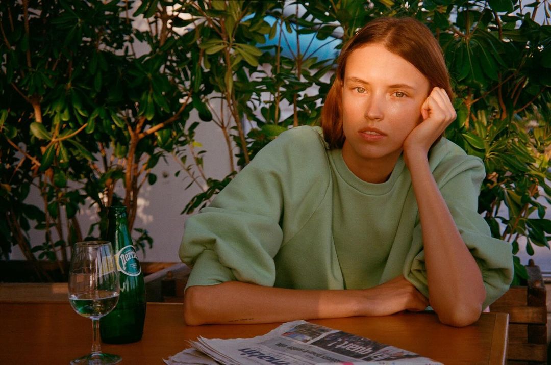 A woman sits at a table with a drink and newspaper, bathed in natural light against a backdrop of leafy greenery.