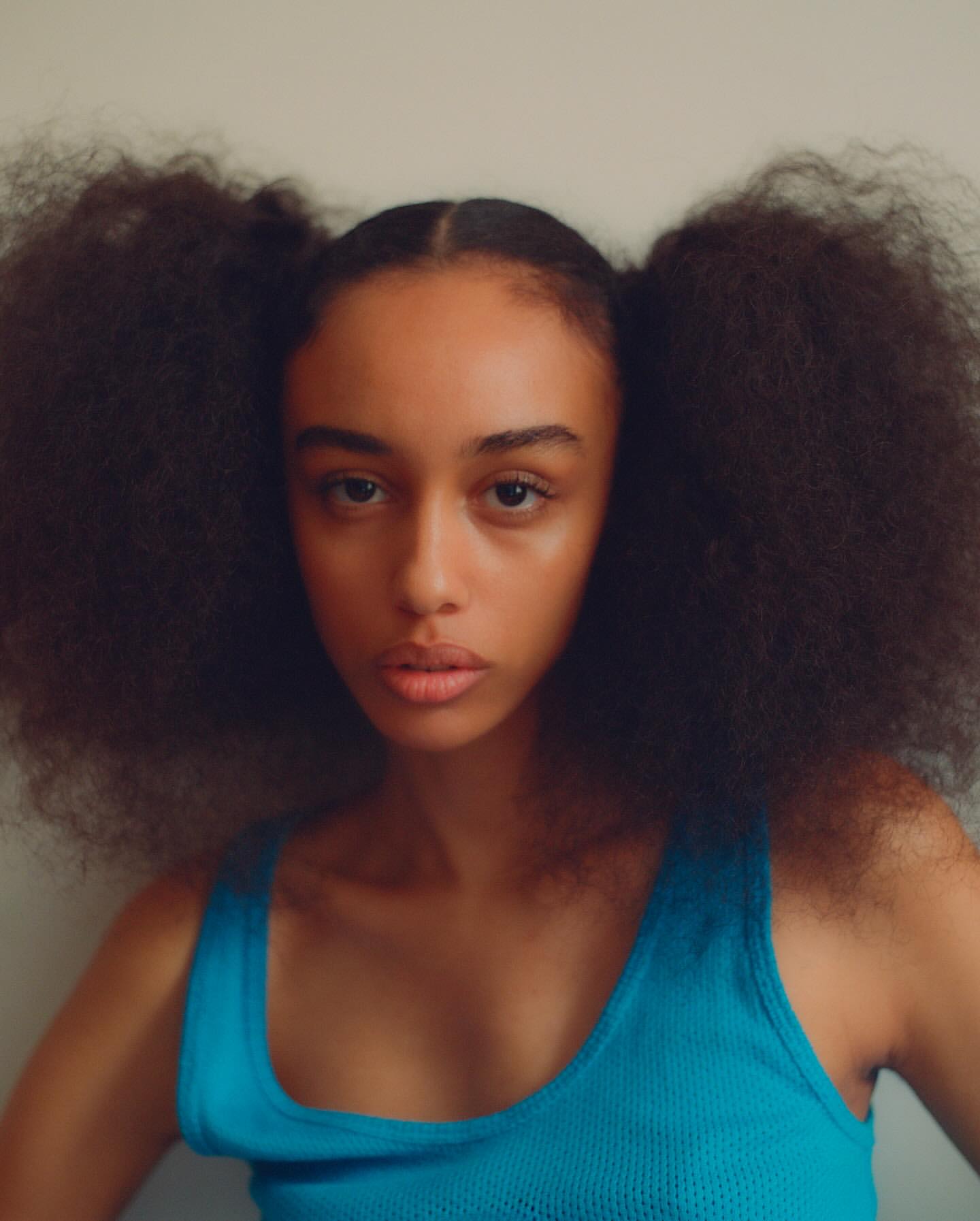 Close-up portrait of a young adult woman with afro pigtails wearing a blue top against a neutral background in a studio setting.