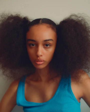 Close-up portrait of a young adult woman with afro pigtails wearing a blue top against a neutral background in a studio setting.