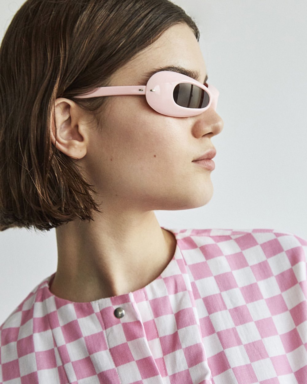 A young adult woman models pink-framed sunglasses and a pink-and-white checkered top against a neutral background in a medium shot.