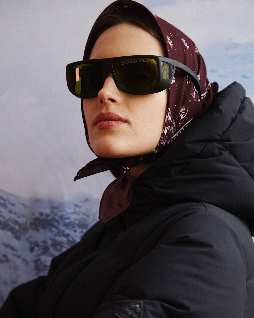 Portrait of a woman wearing sunglasses, a headscarf, and a puffer jacket against a blurred mountain backdrop, evoking winter fashion.
