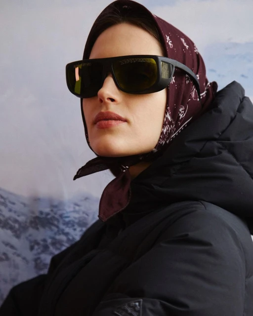 Portrait of a woman wearing sunglasses, a headscarf, and a puffer jacket against a blurred mountain backdrop, evoking winter fashion.