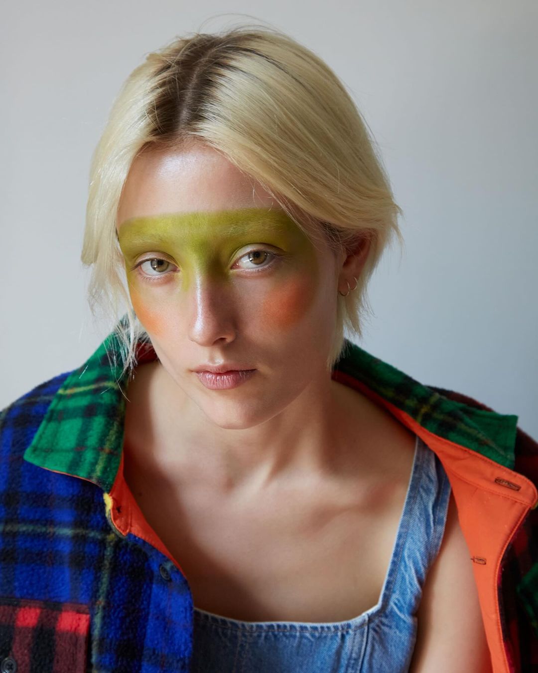 Portrait of a woman with blonde hair and creative makeup including green eyeshadow, orange blush, wearing a denim top and flannel shirt.
