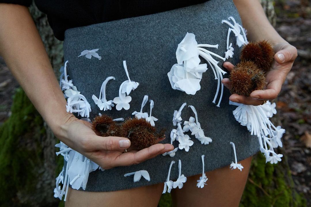 Close-up of a gray skirt decorated with white flowers, held by a woman, with spiky brown seed pods in her hands in a forest setting.