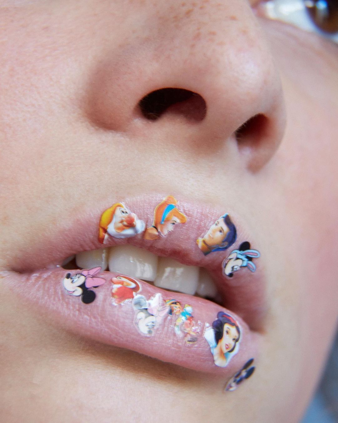 Close-up of a person's face with cartoon character stickers on their lips, including Disney characters such as Snow White and Mickey Mouse.