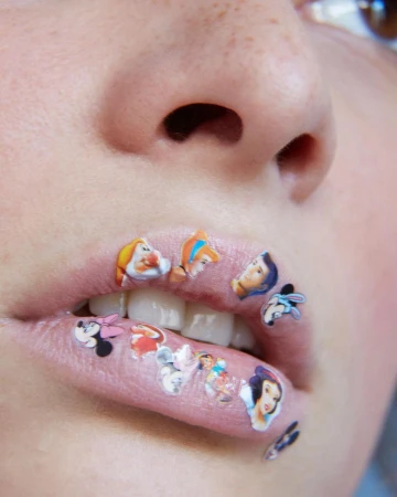 Close-up of a person's face with cartoon character stickers on their lips, including Disney characters such as Snow White and Mickey Mouse.