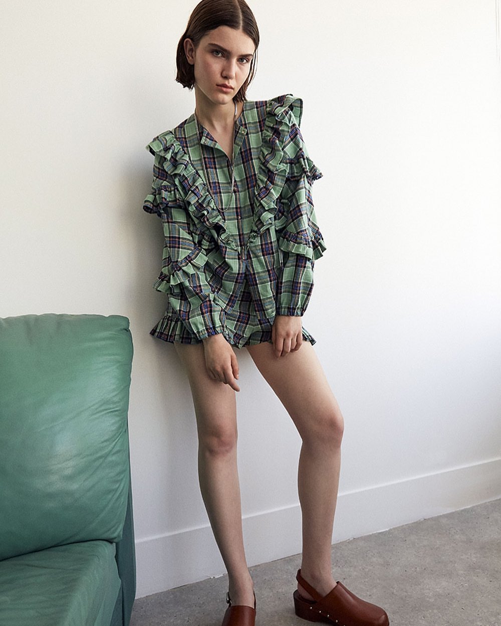A young adult woman poses indoors wearing a ruffled, light green, plaid romper and brown clogs, standing in front of a white wall.