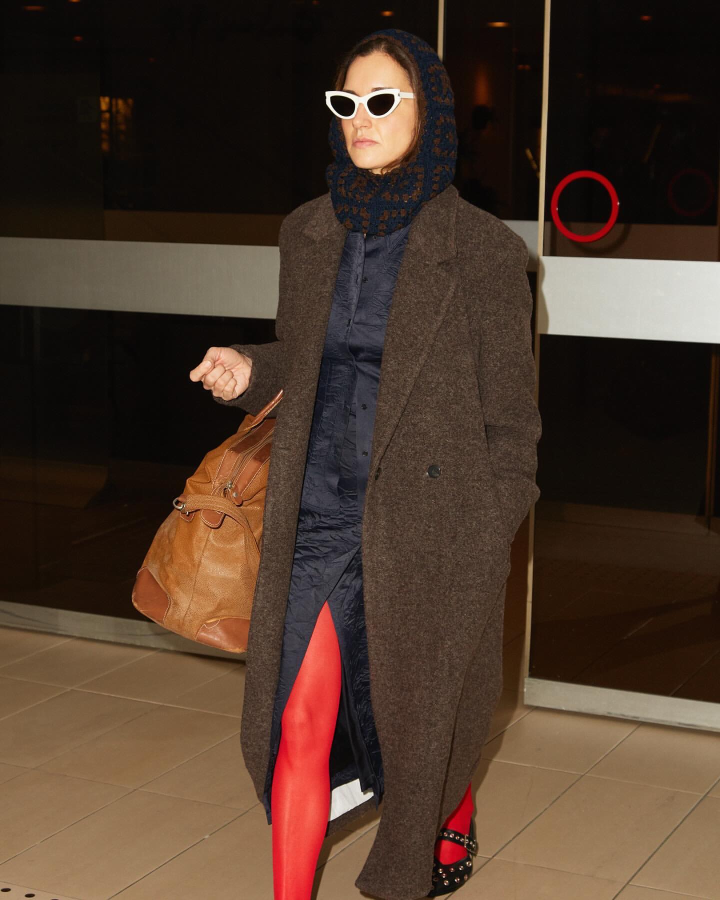 Woman in brown coat, blue dress, red tights and white sunglasses carries a duffel bag, walking in a brightly lit hallway.