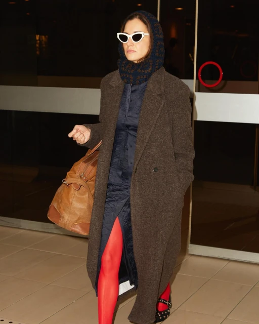 Woman in brown coat, blue dress, red tights and white sunglasses carries a duffel bag, walking in a brightly lit hallway.