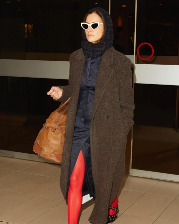 Woman in brown coat, blue dress, red tights and white sunglasses carries a duffel bag, walking in a brightly lit hallway.