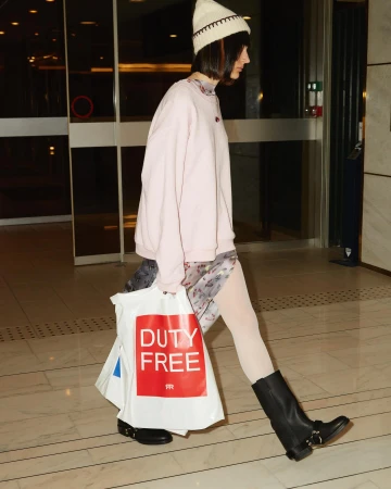 A young adult woman walks indoors with shopping bags, wearing a light pink sweatshirt, patterned dress, and black boots, near glass doors.