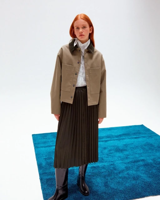 Adult woman in a studio wearing a cropped jacket, pleated skirt, shirt, and knee-high boots standing on a blue textured rug.