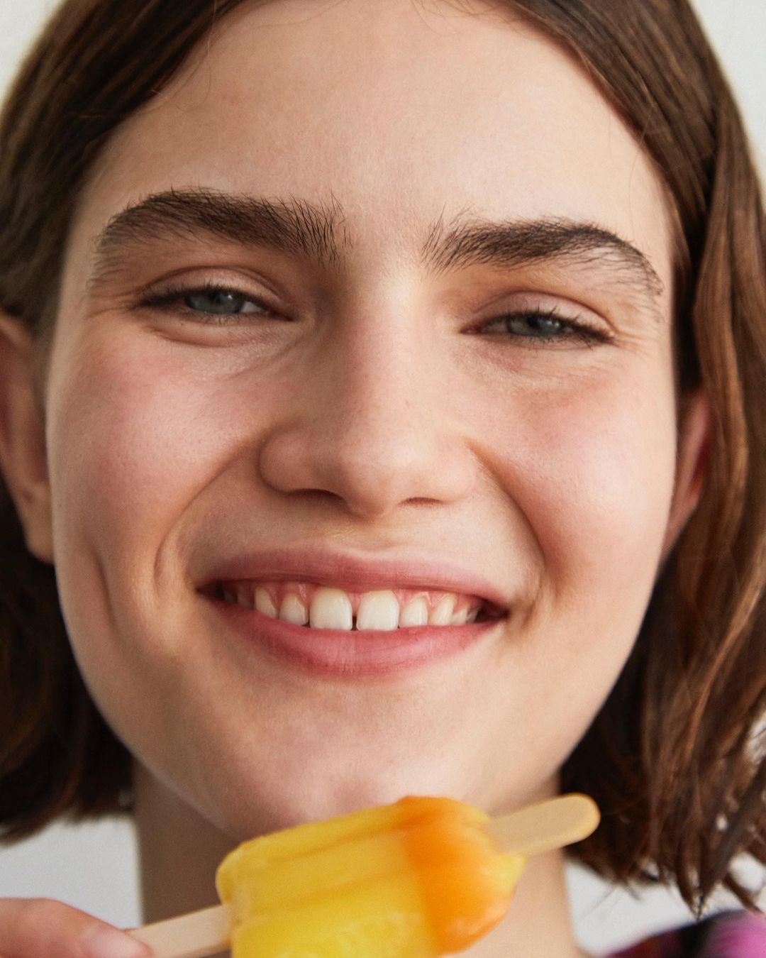 Close-up portrait of a smiling woman holding a yellow and orange popsicle, showcasing youthful beauty and a refreshing summer treat.