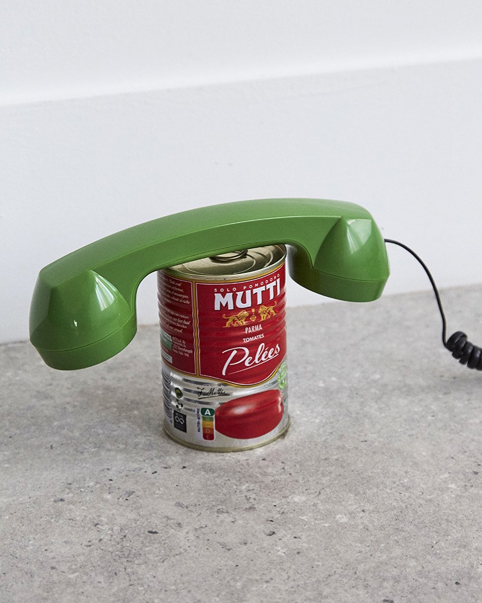 Still life shot of a green vintage telephone handset resting on top of a can of Mutti brand peeled tomatoes on a grey surface.