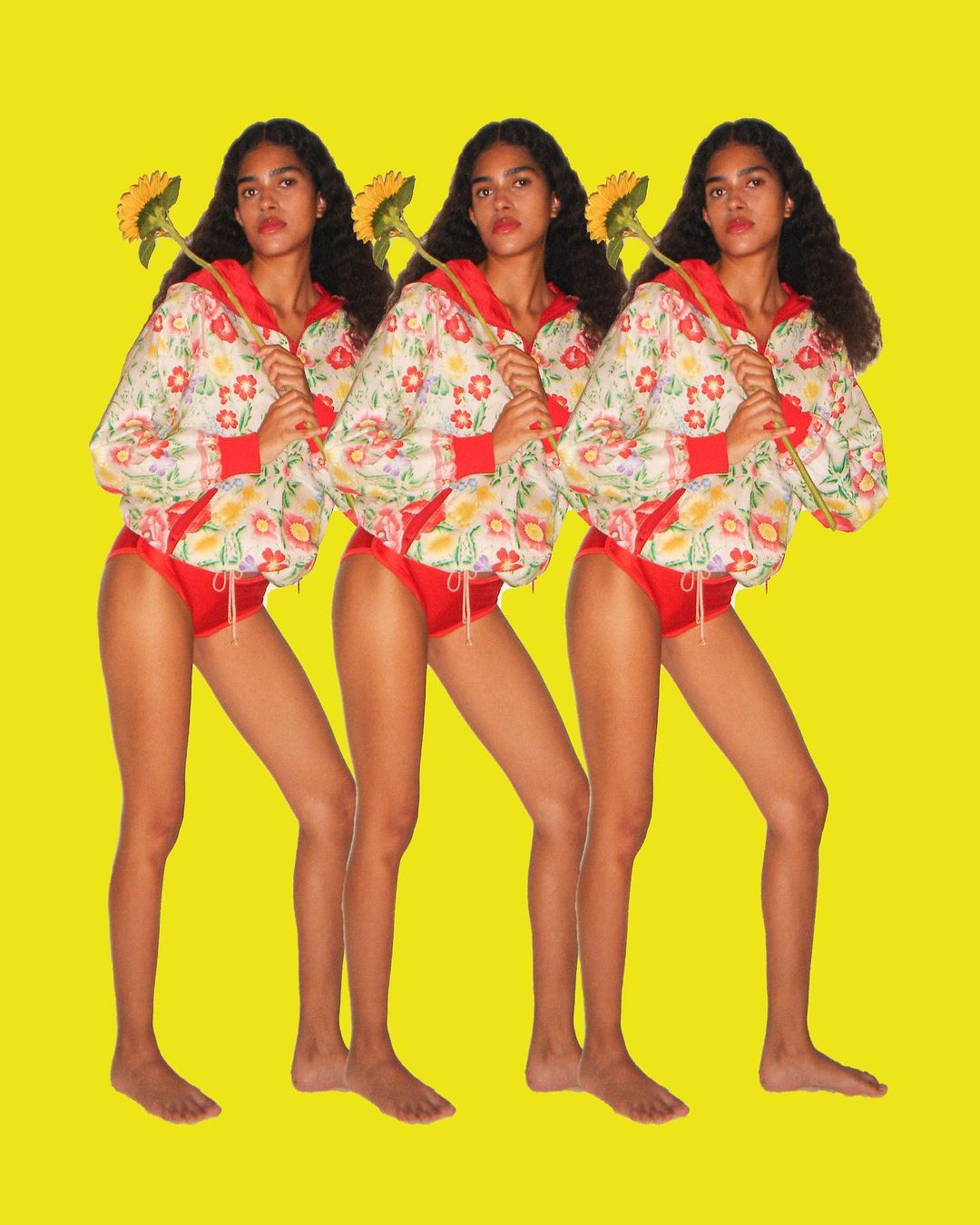 Three young women stand against a yellow backdrop, each holding a sunflower, wearing a floral jacket and red bottoms in a studio setting.