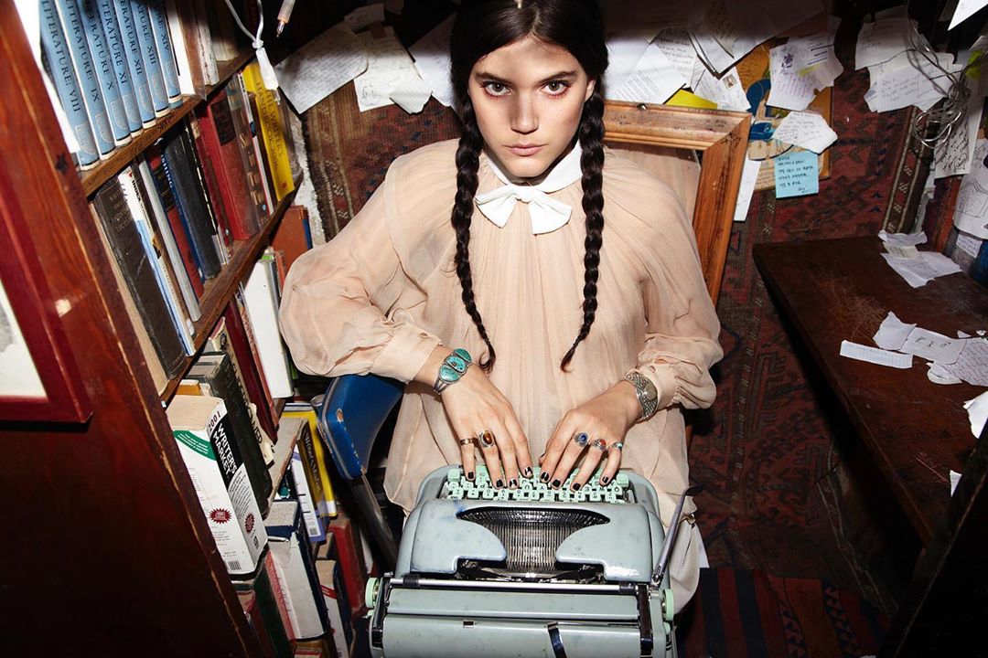 Woman types on vintage typewriter in book-filled room. She wears light-colored blouse with collar bow and braided hair, rings, bracelets.