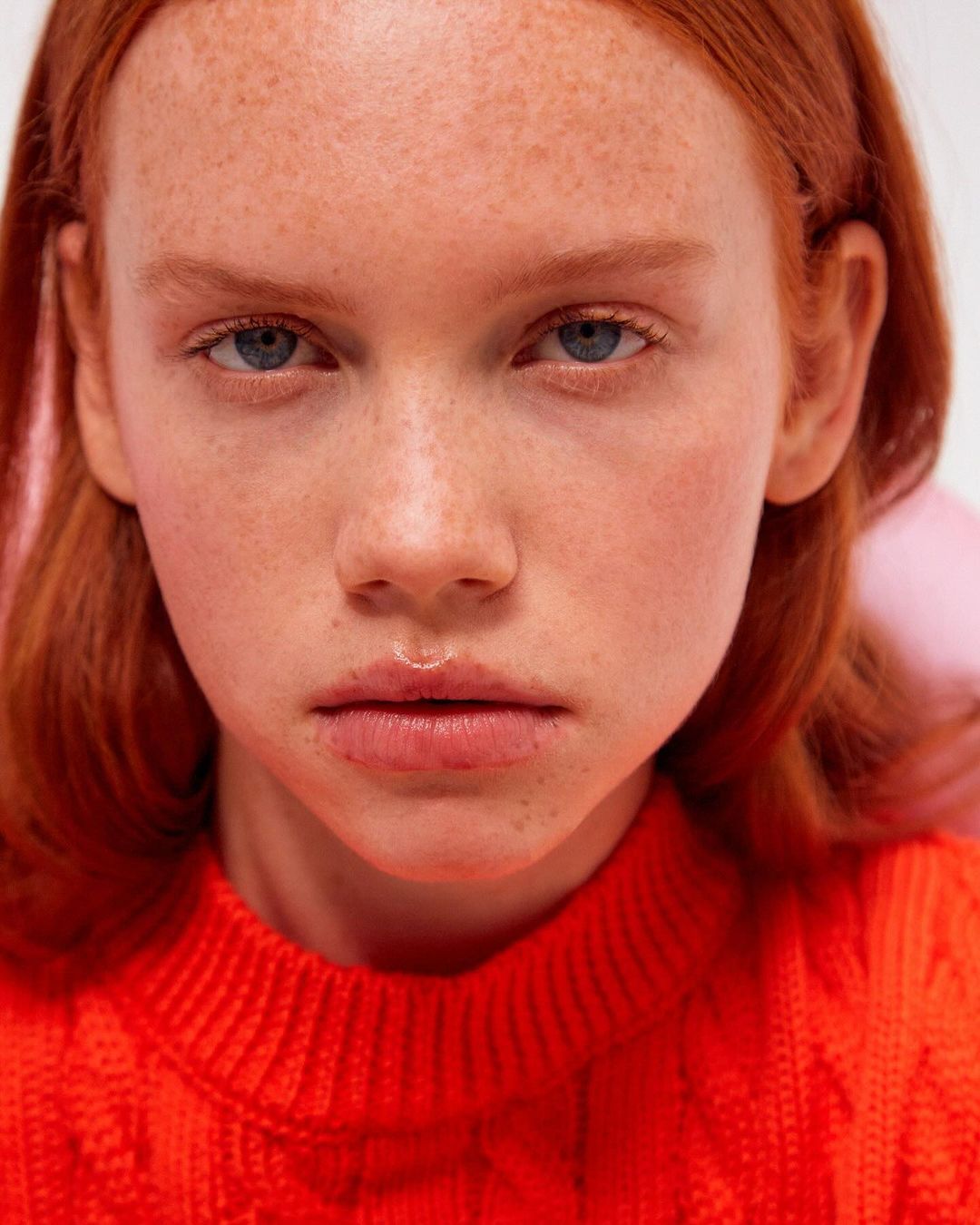 Close-up portrait of a red-haired woman with freckles wearing a vibrant orange knitted sweater against a plain background