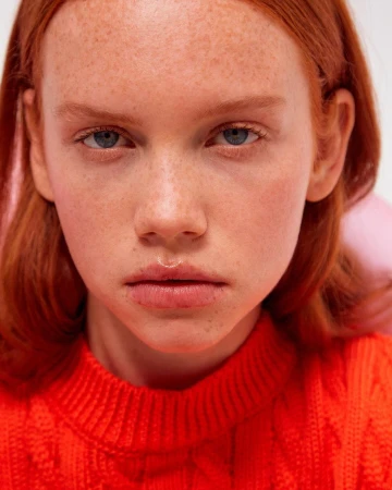 Close-up portrait of a red-haired woman with freckles wearing a vibrant orange knitted sweater against a plain background