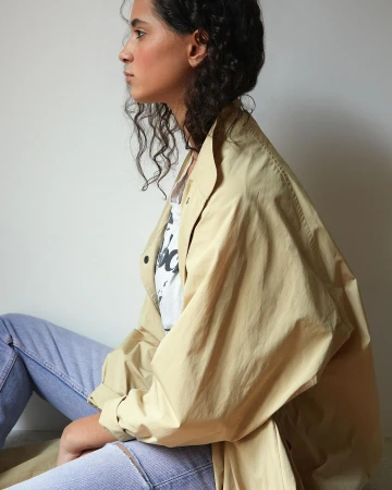 Profile portrait of a woman wearing a beige trench coat and light blue jeans against a plain wall.