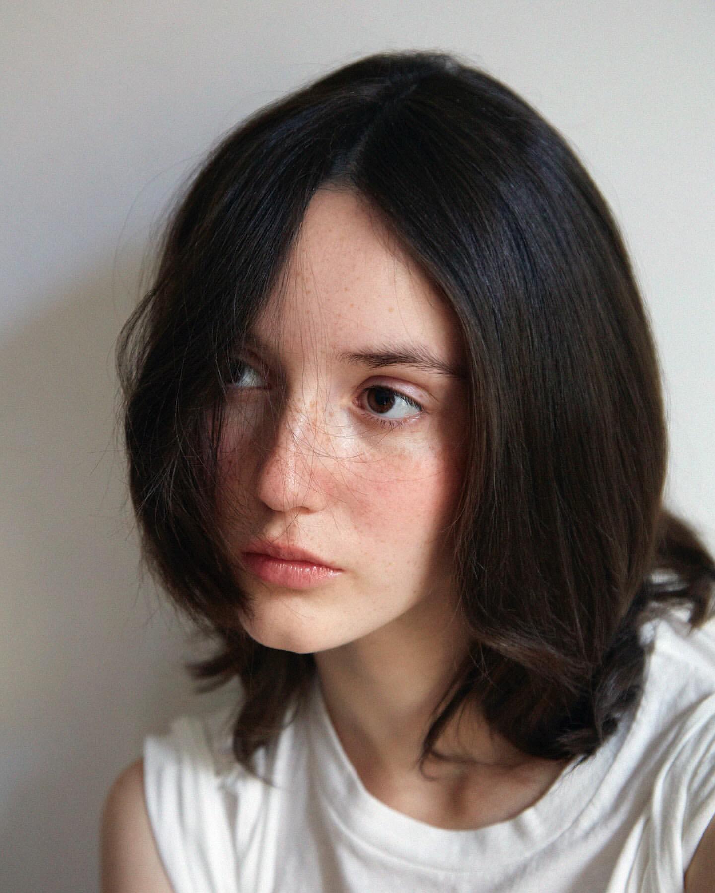 Portrait of a young woman with dark hair and freckles wearing a white t-shirt against a neutral background