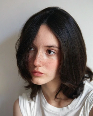 Portrait of a young woman with dark hair and freckles wearing a white t-shirt against a neutral background