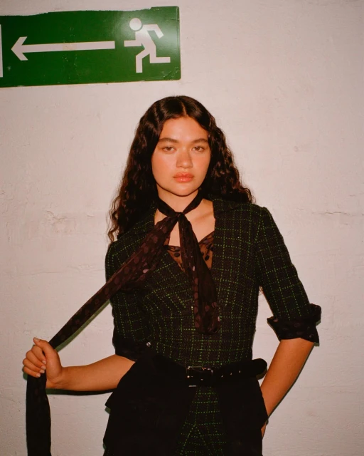 Portrait of a young woman with dark wavy hair wearing a blazer with a polka dot bow standing in front of a green exit sign.