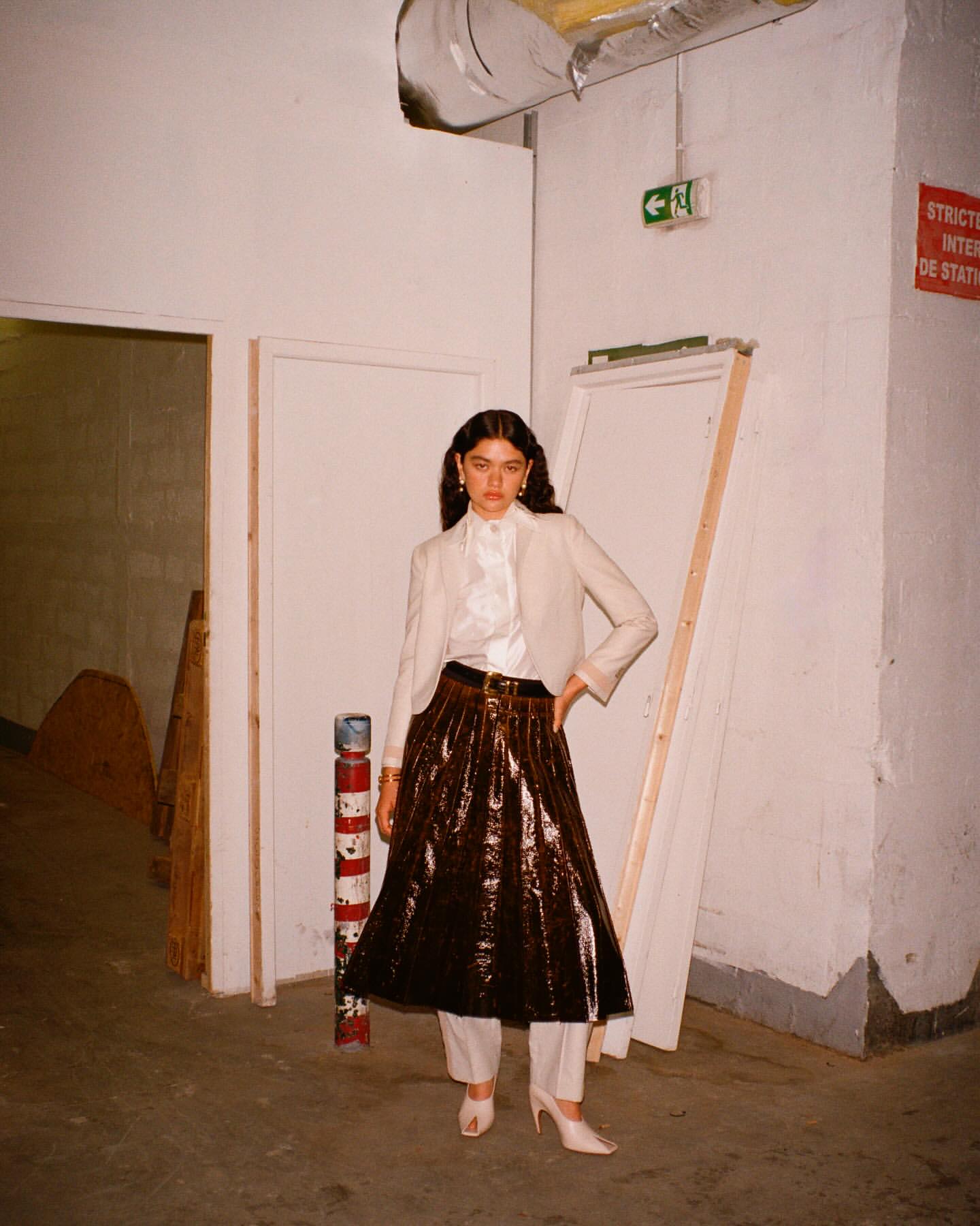 Woman poses in a beige blazer, pleated brown skirt, and white heels in an urban, indoor setting with white walls and industrial details.