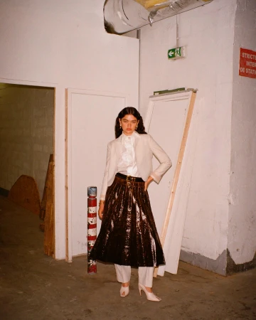 Woman poses in a beige blazer, pleated brown skirt, and white heels in an urban, indoor setting with white walls and industrial details.