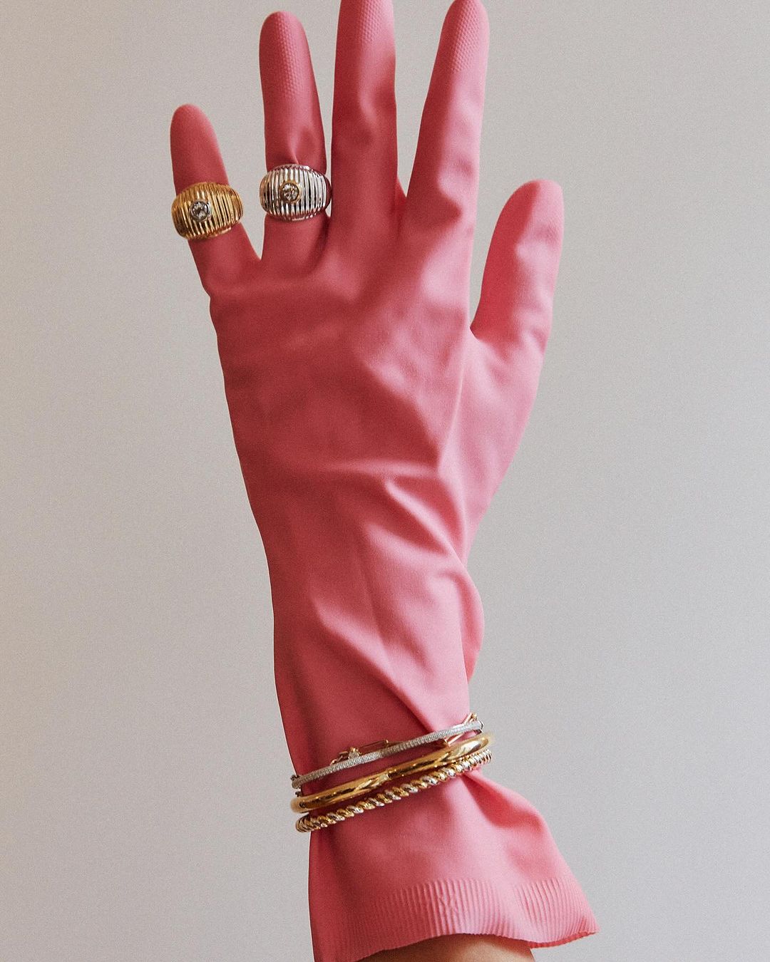 Hand wearing a pink rubber glove accessorized with silver and gold rings and bracelets against a white background in a vertical shot.