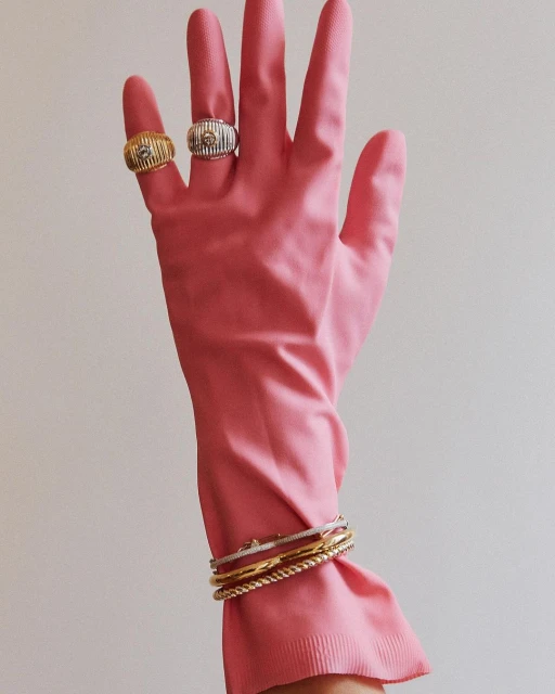 Hand wearing a pink rubber glove accessorized with silver and gold rings and bracelets against a white background in a vertical shot.
