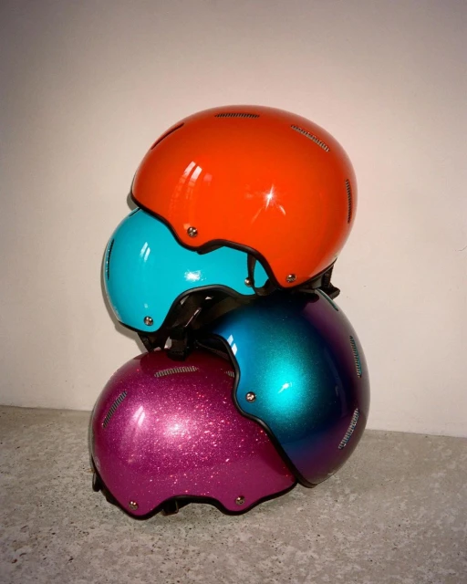 Stack of colorful protective helmets: orange, turquoise, and purple, arranged on a concrete floor against a neutral wall backdrop.